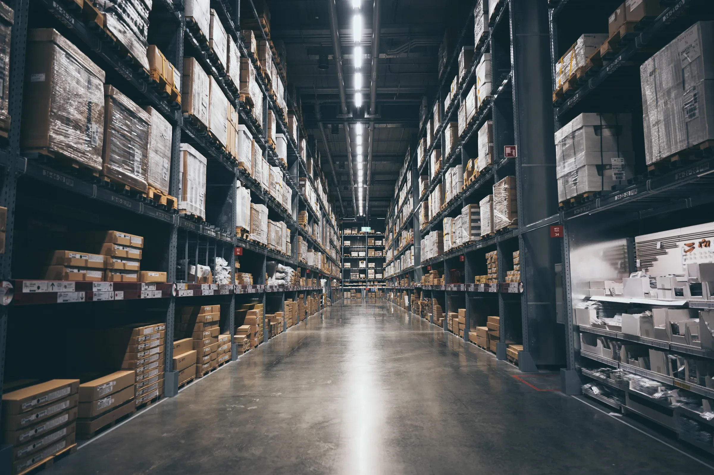 A long warehouse aisle of stocked shelving, lit from the far end